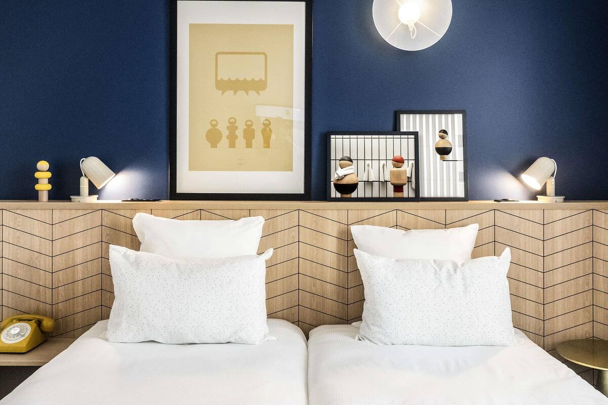 A hotel room with a bed, two pillows, a framed artwork, a wall-mounted light, and a small table with a lamp.