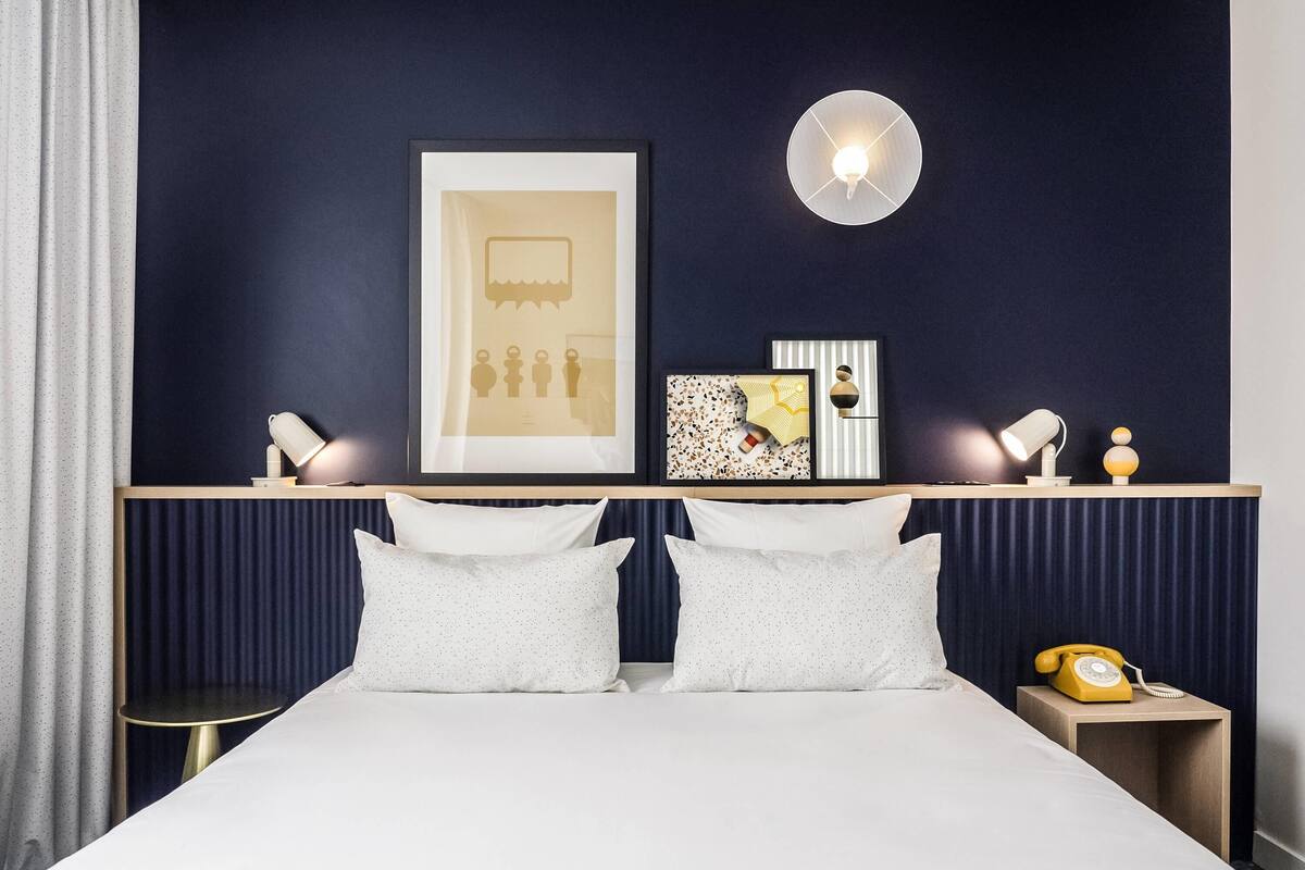 A hotel room with a bed, two bedside tables, a nightstand with a telephone, a wall-mounted light, and a framed picture above the bed.