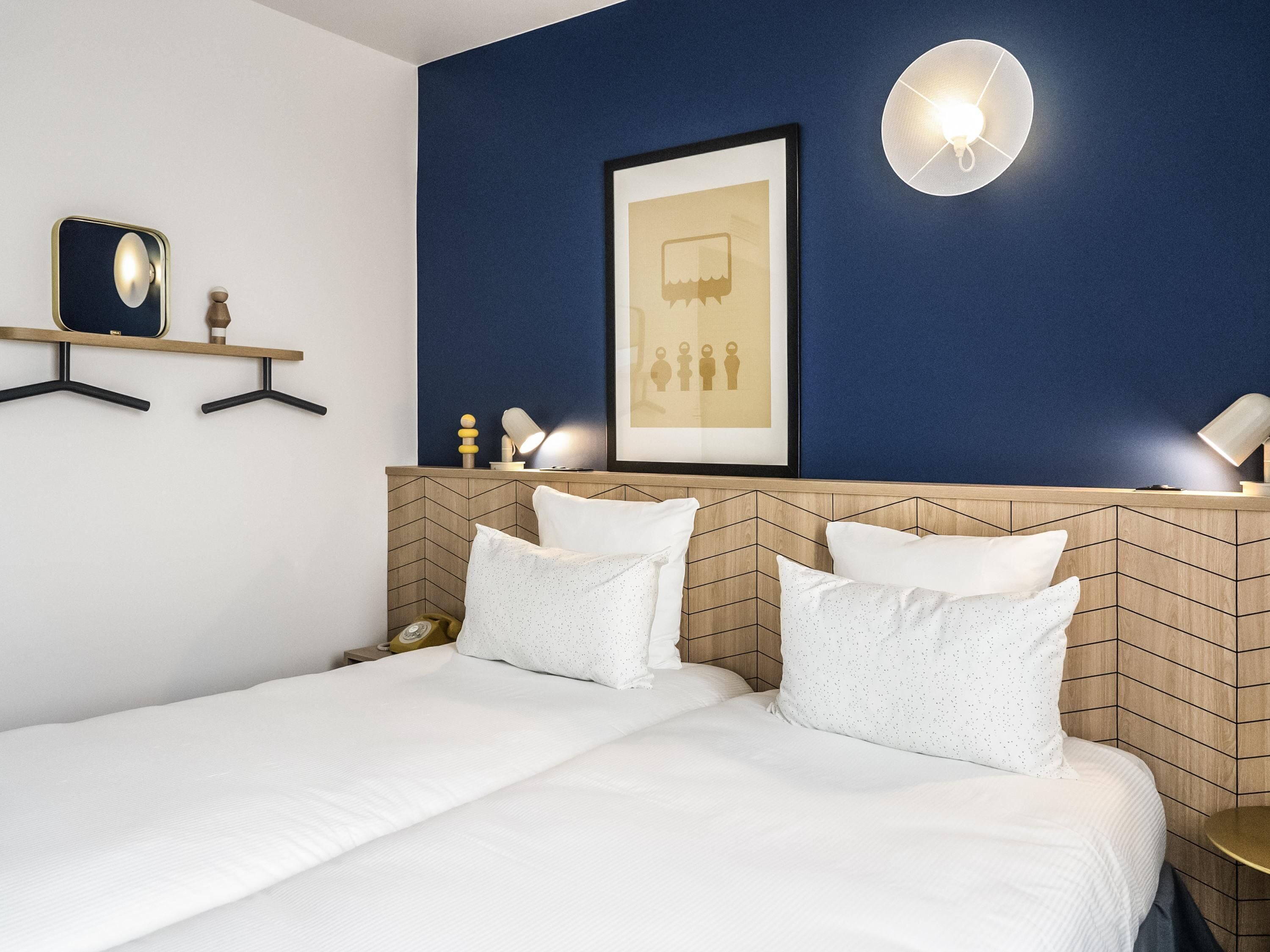 A hotel room with a bed, a headboard with a geometric pattern, a framed picture on the wall, and a wall-mounted light fixture.