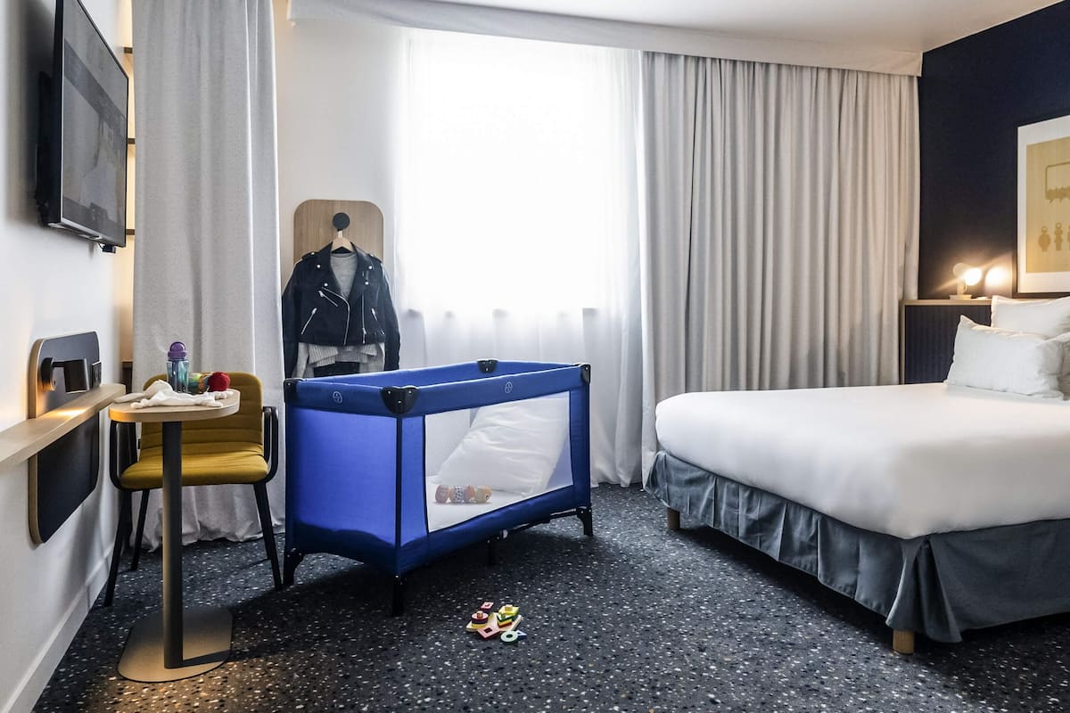 A hotel room with a bed, a desk with a chair, a blue crib, and a window with curtains.