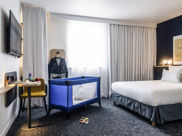 Habitación de hotel con cama, escritorio y silla, una cuna azul y ventana con cortinas.