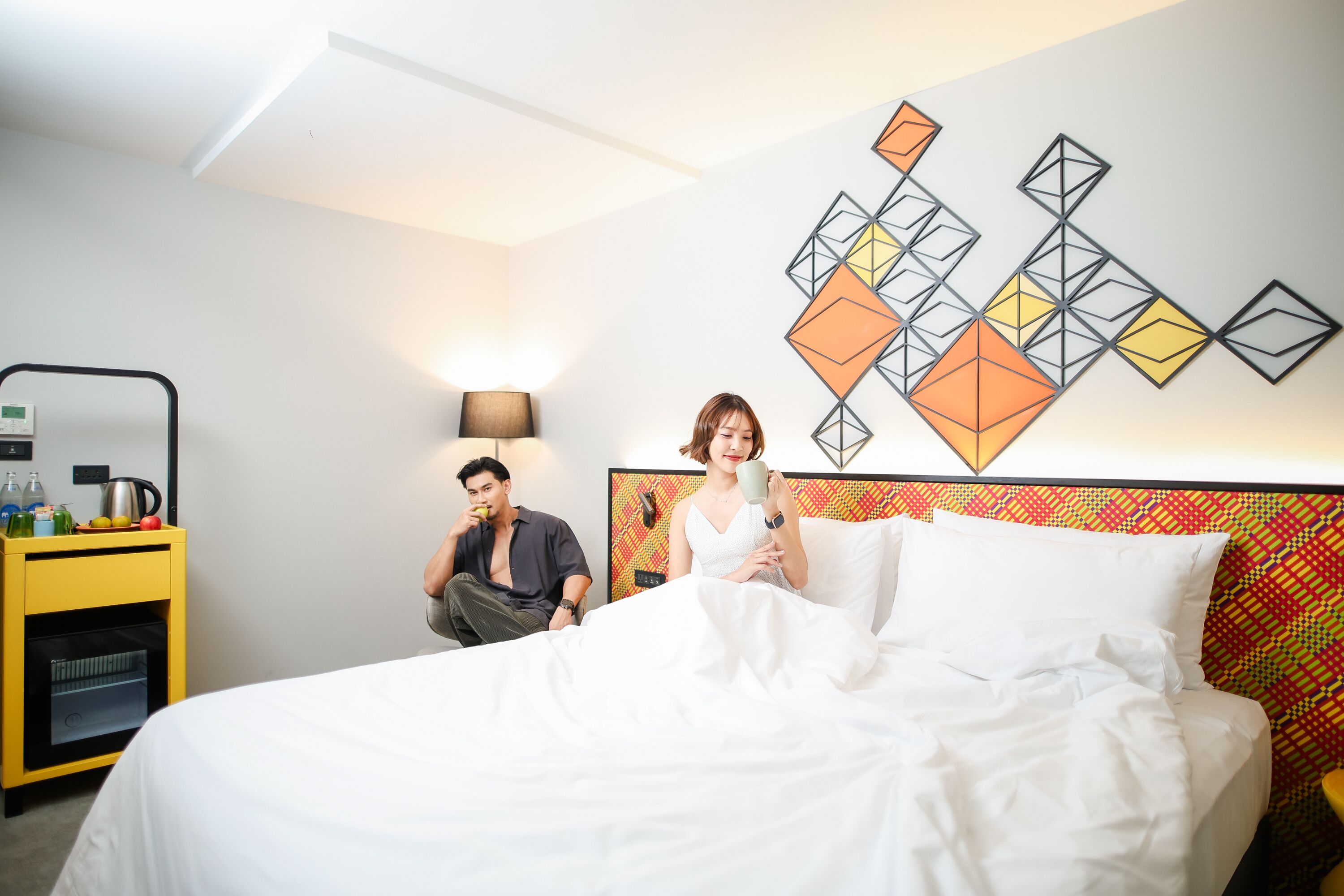 A hotel room with a bed, a bedside table with a lamp, and a wall art piece.