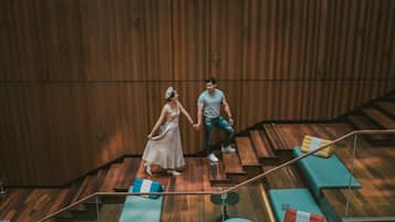 Una pareja tomados de la mano en una escalera de madera con cojines verdes y rojos, una pequeña mesa redonda y una barandilla de vidrio.