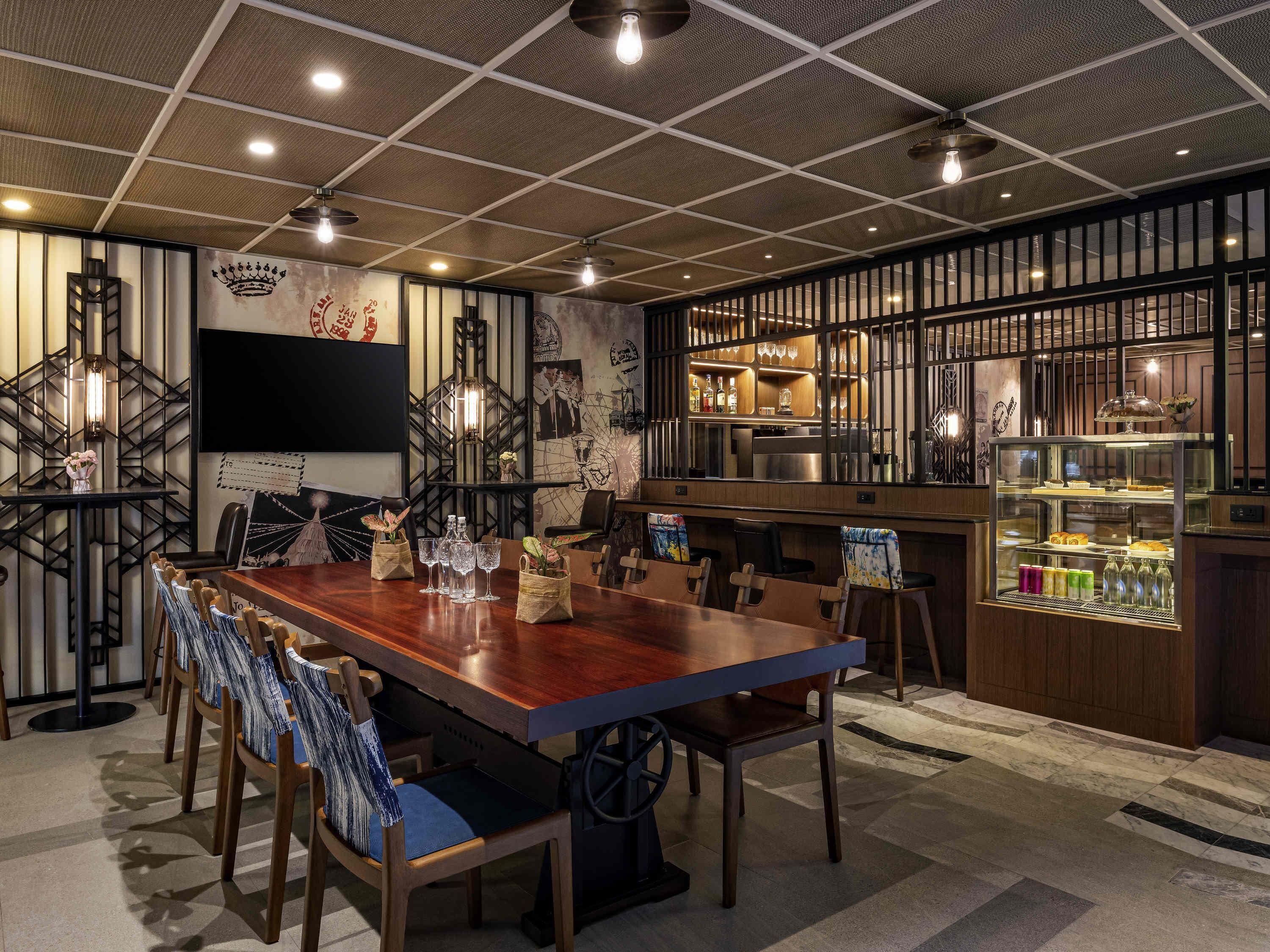 Un restaurante con una larga mesa de madera, sillas, una zona de bar y un televisor en la pared.