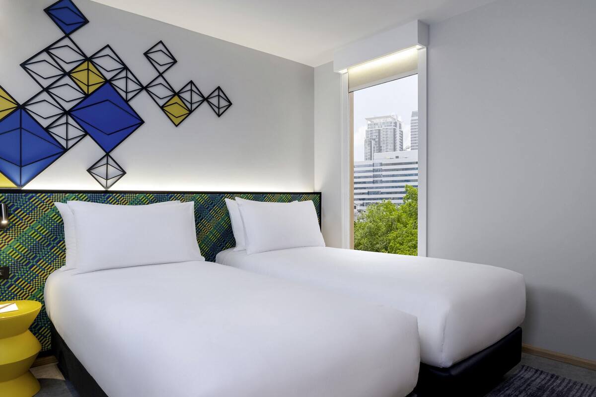 A hotel room with two beds, a patterned wall, a window with a city view, and a yellow side table.