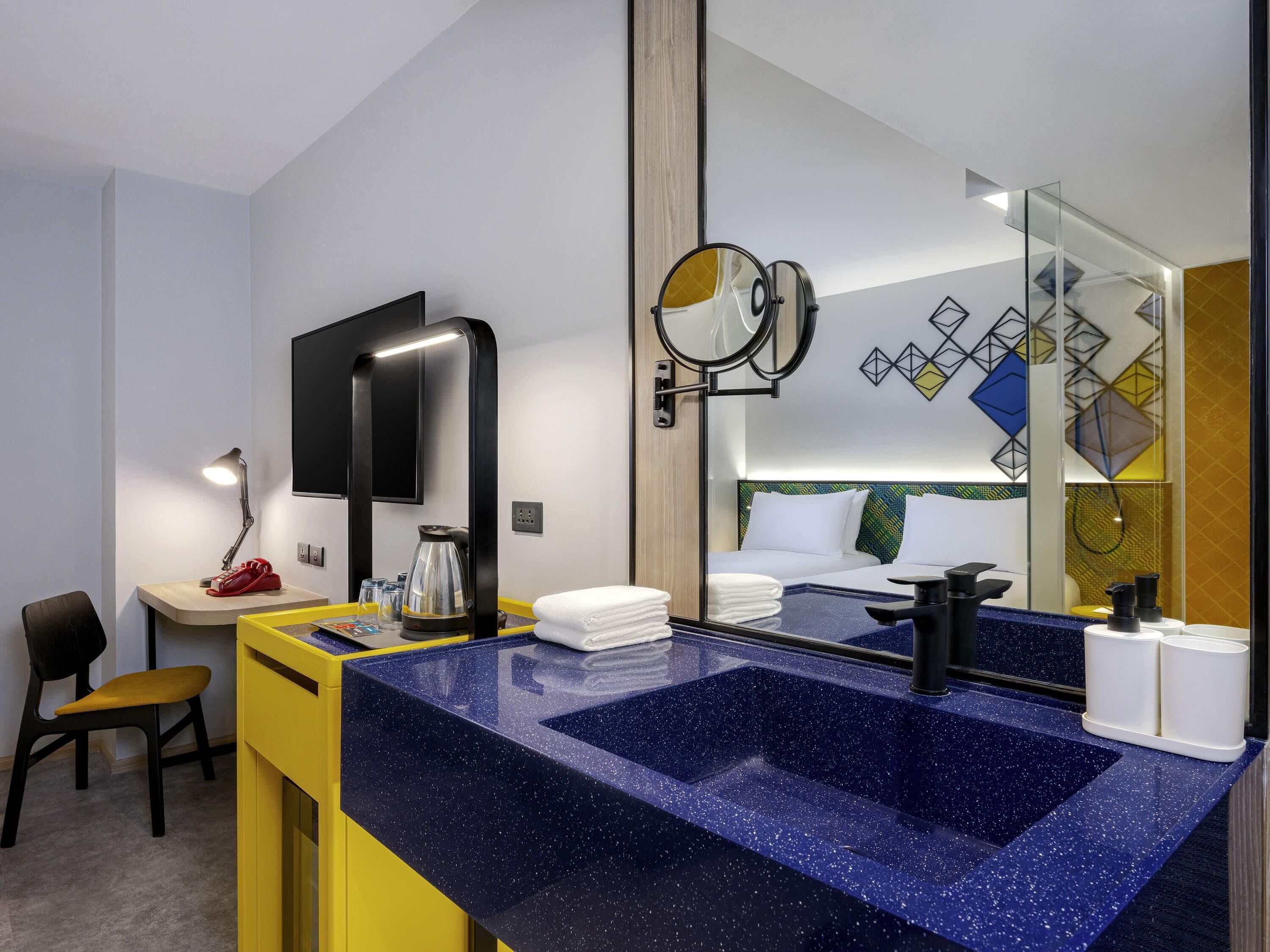 A hotel room with a large mirror, a desk with a lamp, a coffee maker, and a blue speckled sink.