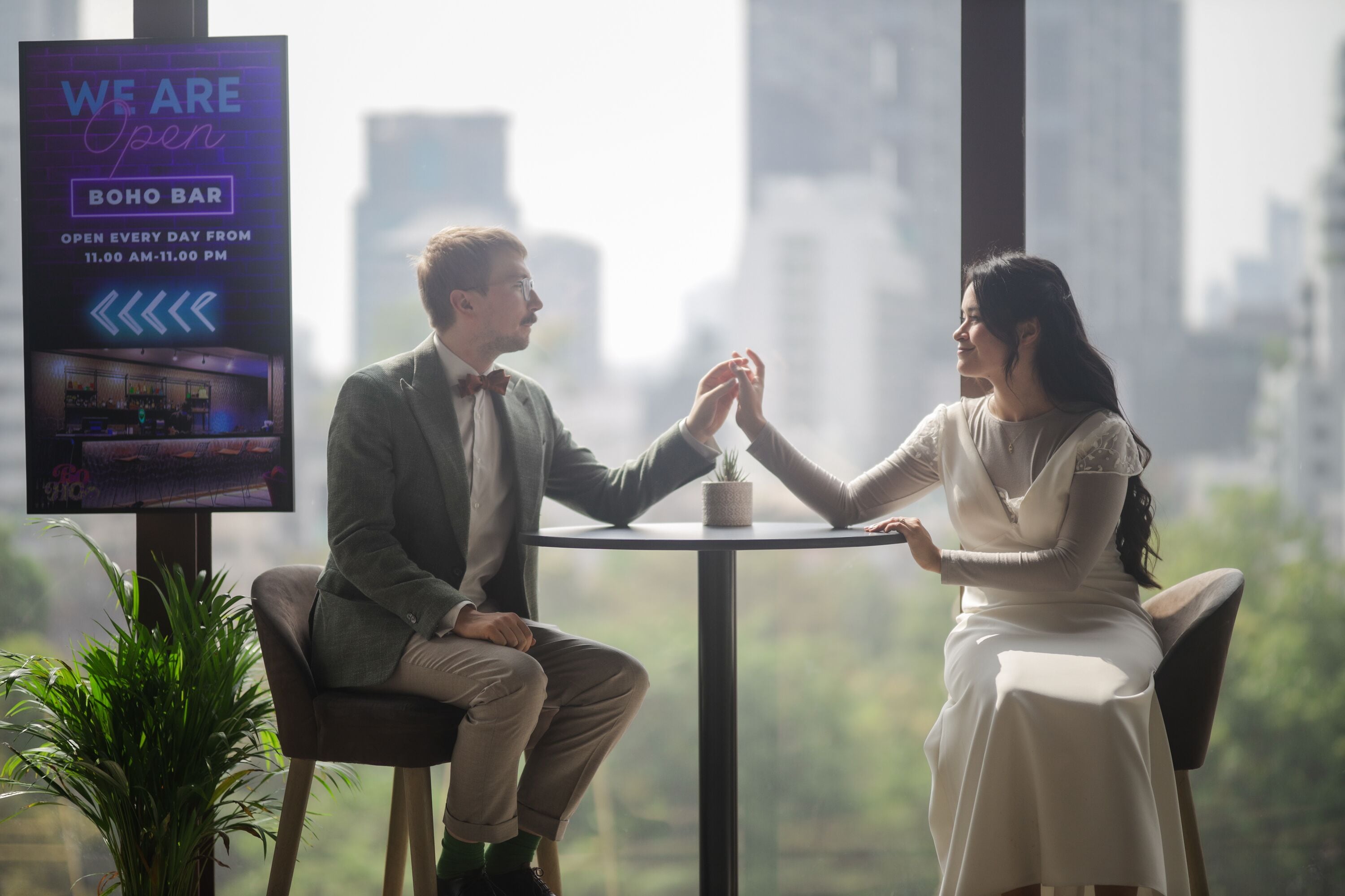 Zwei Personen sitzen an einem Tisch in einer modernen Bar mit Blick auf die Stadt, einer Neonreklame und einem Topfpflanzen.