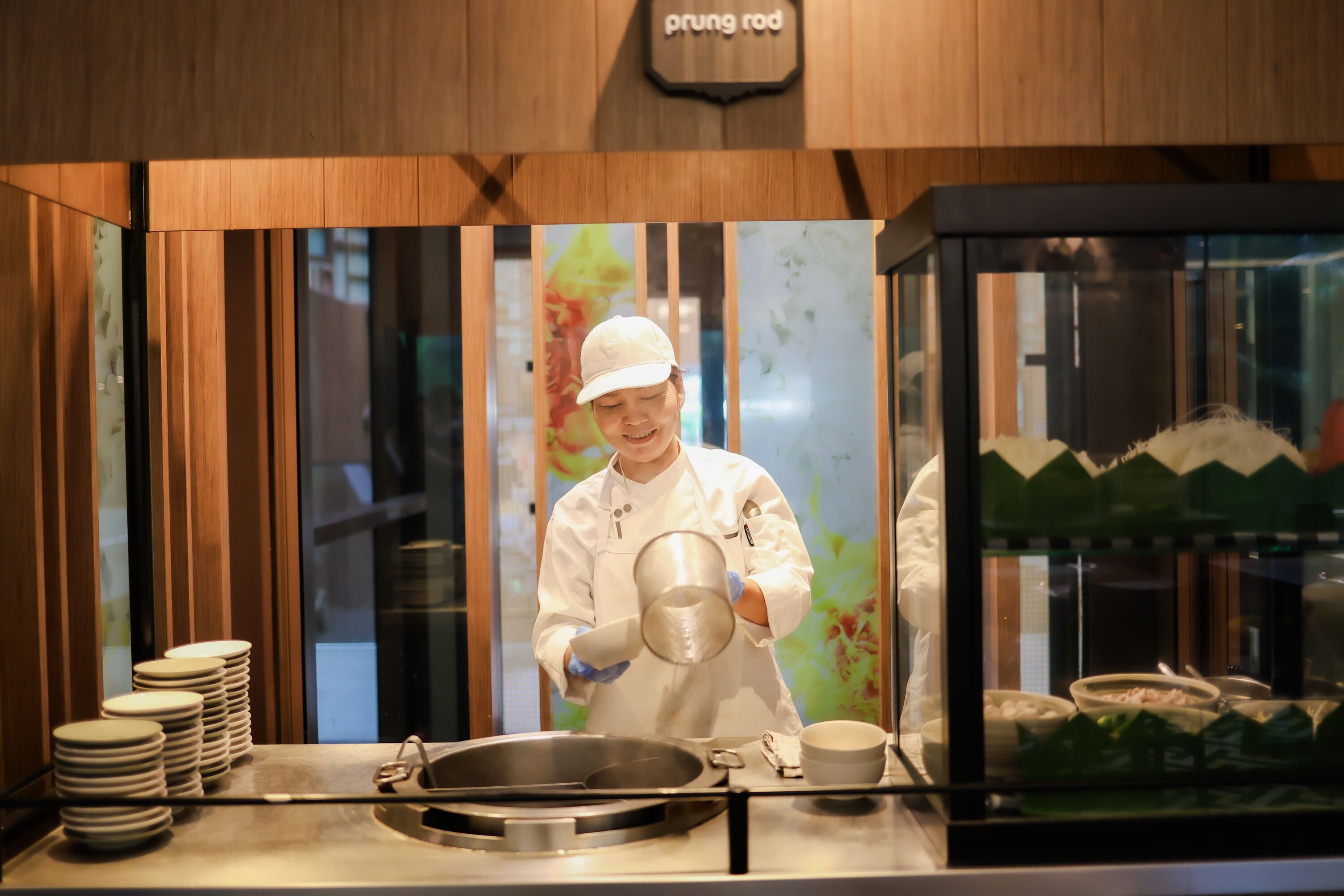 A chef in a white uniform pouring liquid into a pan on a stainless steel countertop.