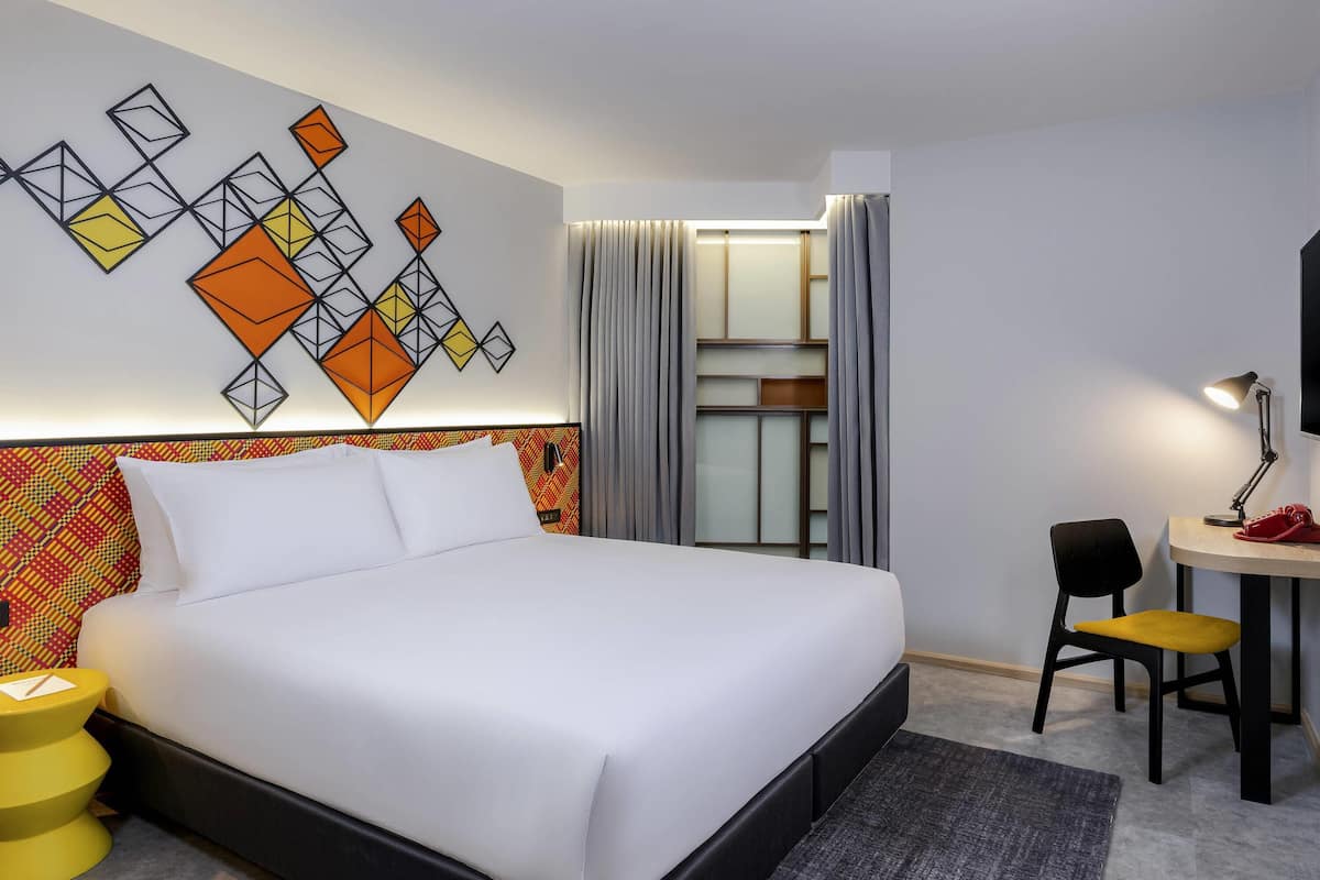 A hotel room with a bed, a desk with a chair, a lamp, and a wall art piece.