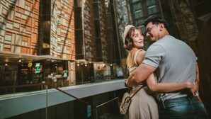Una pareja abrazándose en un edificio moderno con ventanas amplias y barandilla de vidrio.