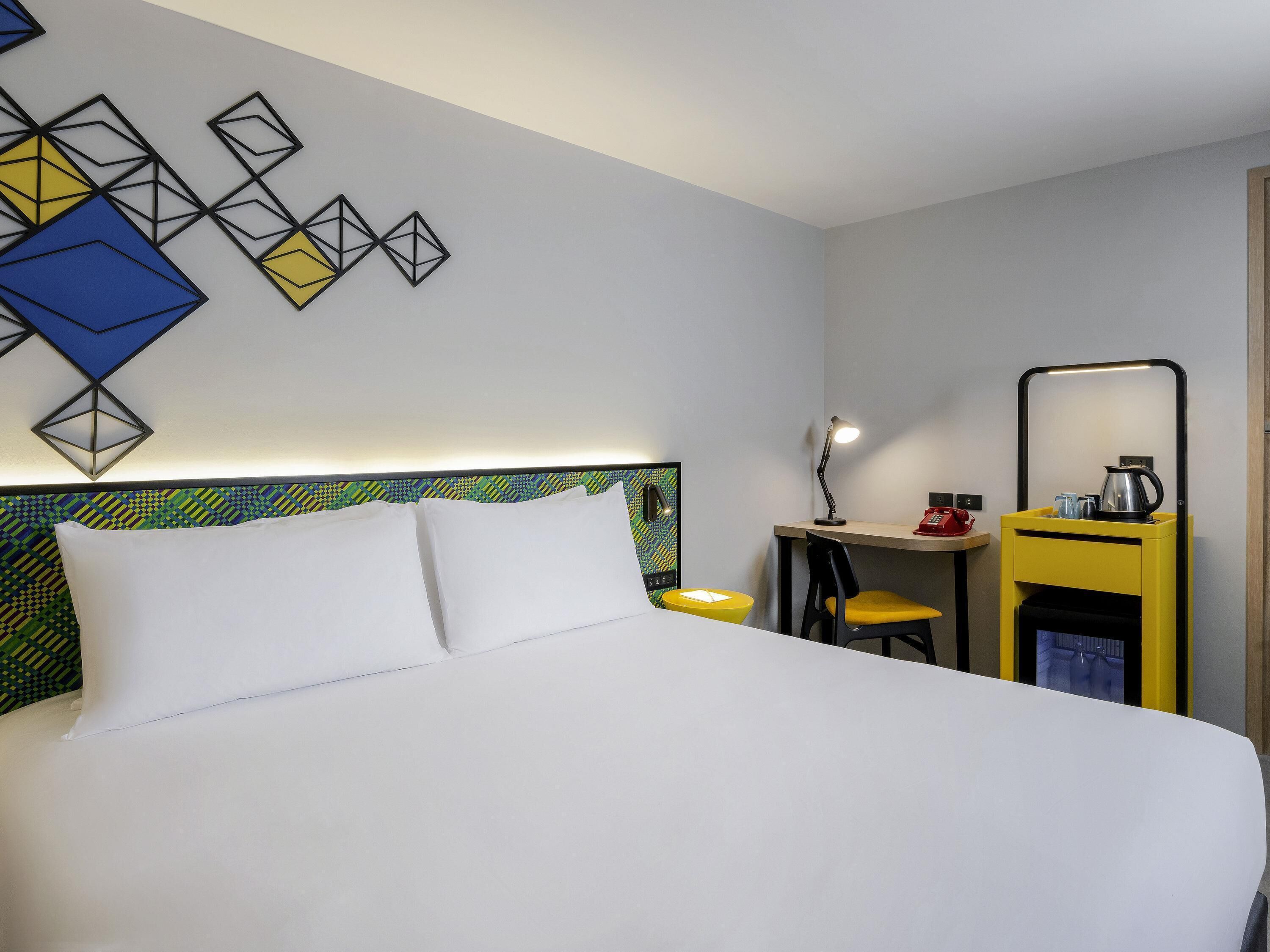 A hotel room with a bed, a desk, a chair, a small table with a kettle, and a mirror.
