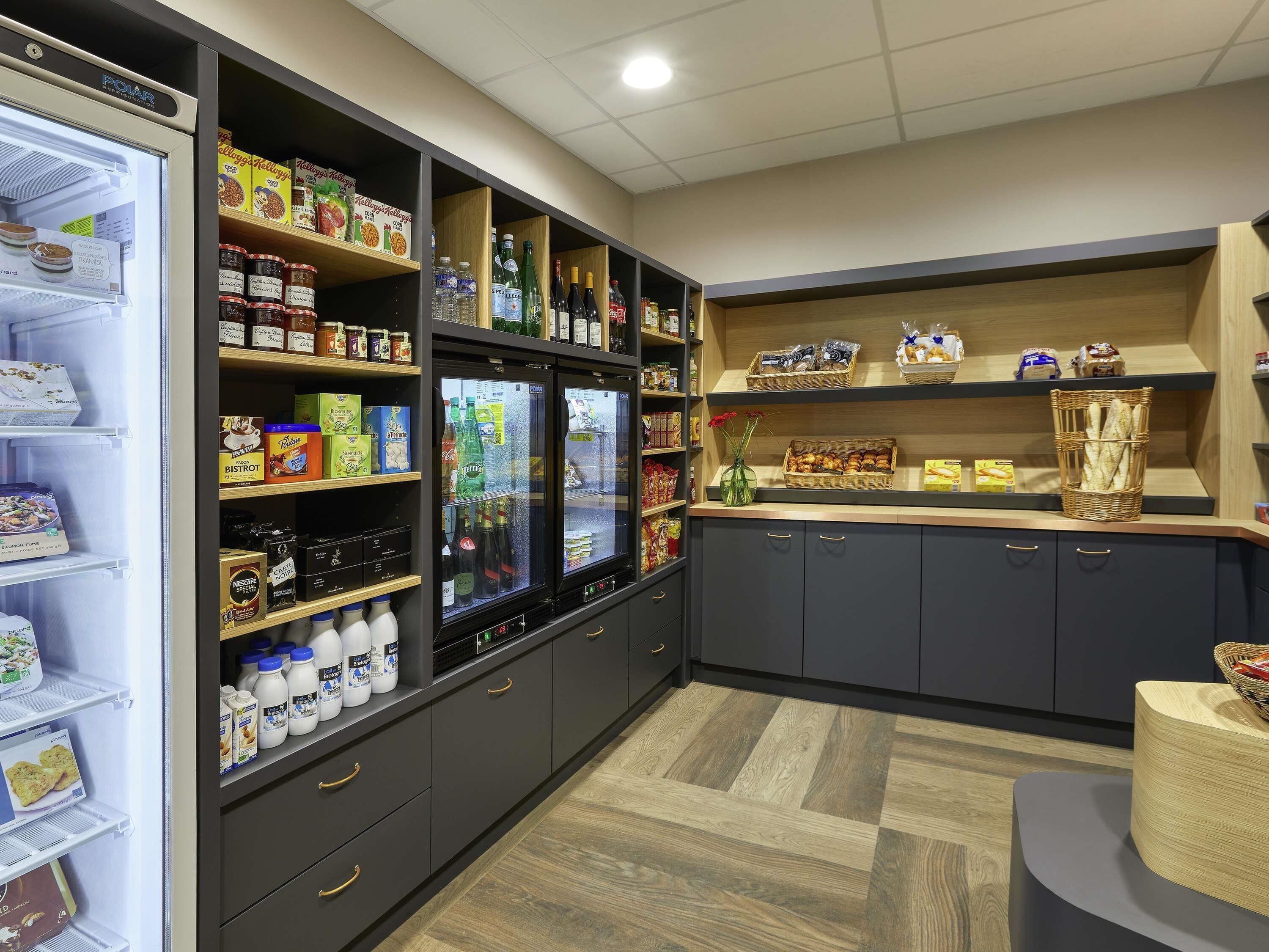A kitchenette with a refrigerator, shelves stocked with food items, and a counter with a basket of bread.