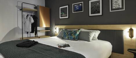A hotel room with a bed, a bedside table, a lamp, a wall-mounted shelf with clothes, and framed pictures on the wall.