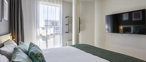 Habitación de hotel con una cama grande, un televisor de pantalla plana en la pared, una ventana con cortinas translúcidas y vistas al exterior.