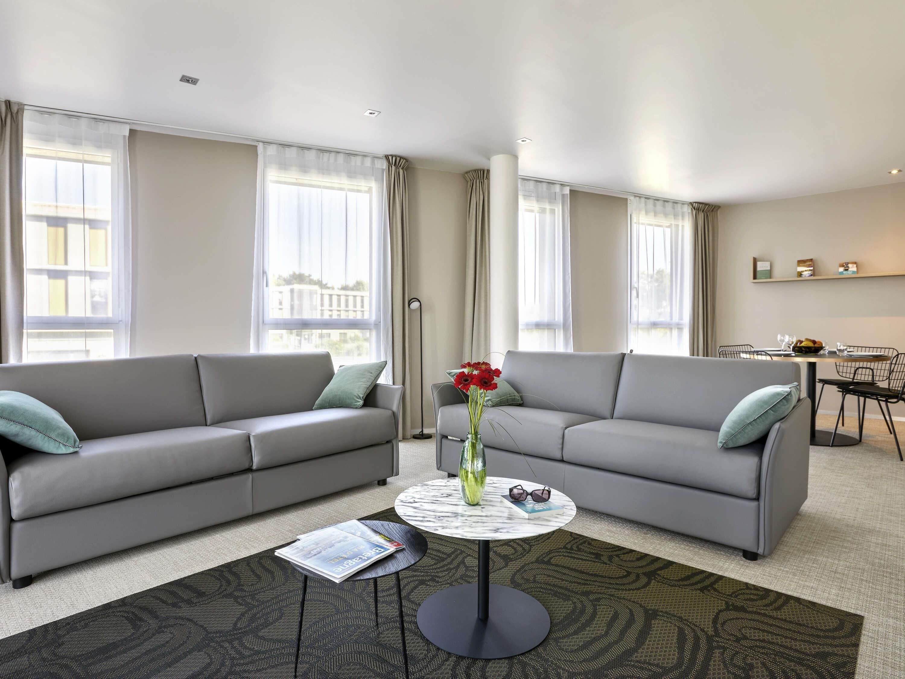 A modern living room with two grey sofas, a round coffee table, and a dining area in the background.