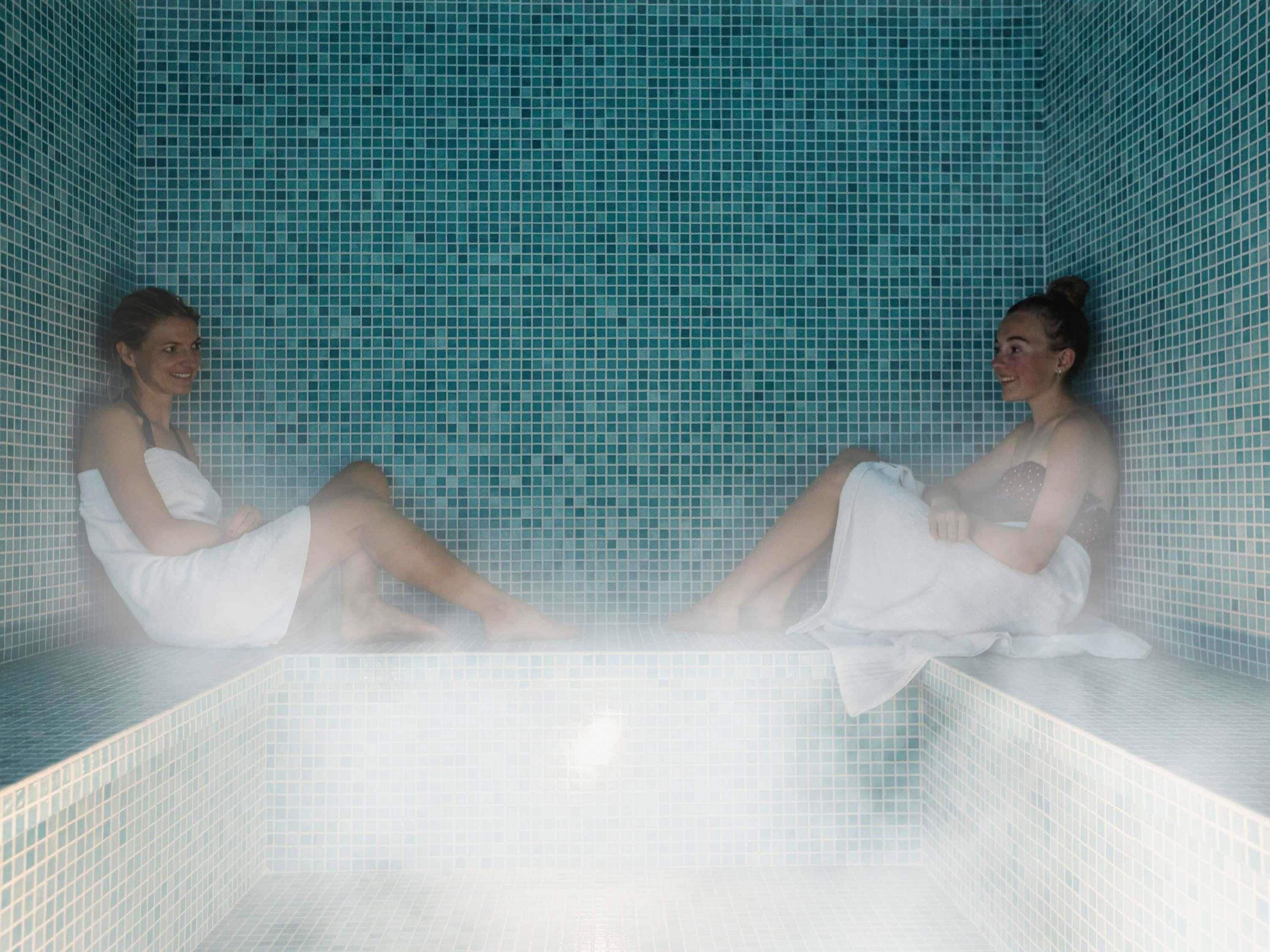 Two individuals seated in a tiled bathtub with a blue mosaic wall.