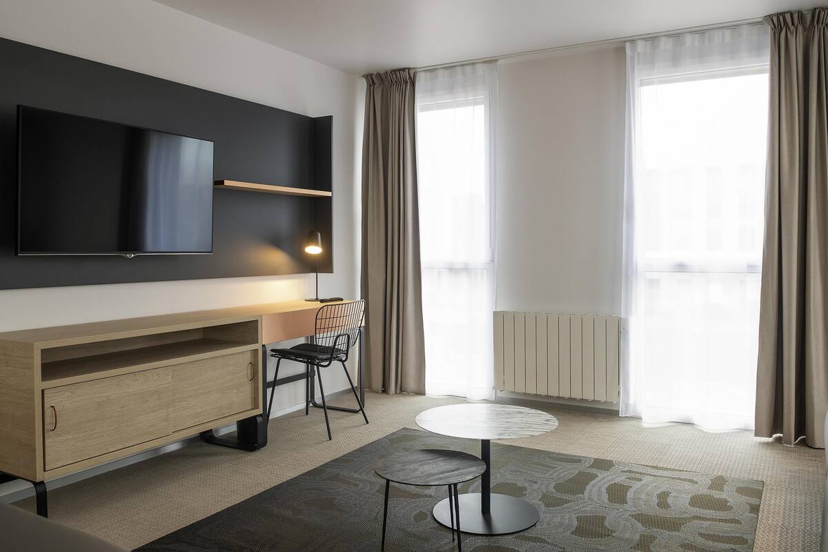 A modern hotel room with a flat-screen TV, a wooden cabinet, a round table, a chair, and large windows with curtains.