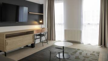 Moderni hotellihuone, jossa on taulutelevisio, puinen kaappi, pyöreä pöytä, tuoli ja suuret ikkunat, joissa on verhot.