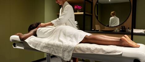 A massage therapy room with a client lying on a treatment table, a therapist in a white robe, and a large mirror on the wall.