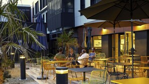 Un área de estar al aire libre con mesas y sillas, rodeada de plantas y una fachada de edificio moderna.