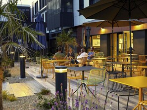 An outdoor seating area with tables and chairs, surrounded by plants and a modern building facade. - Aparthotel Adagio & Spa Vannes (Plescop)