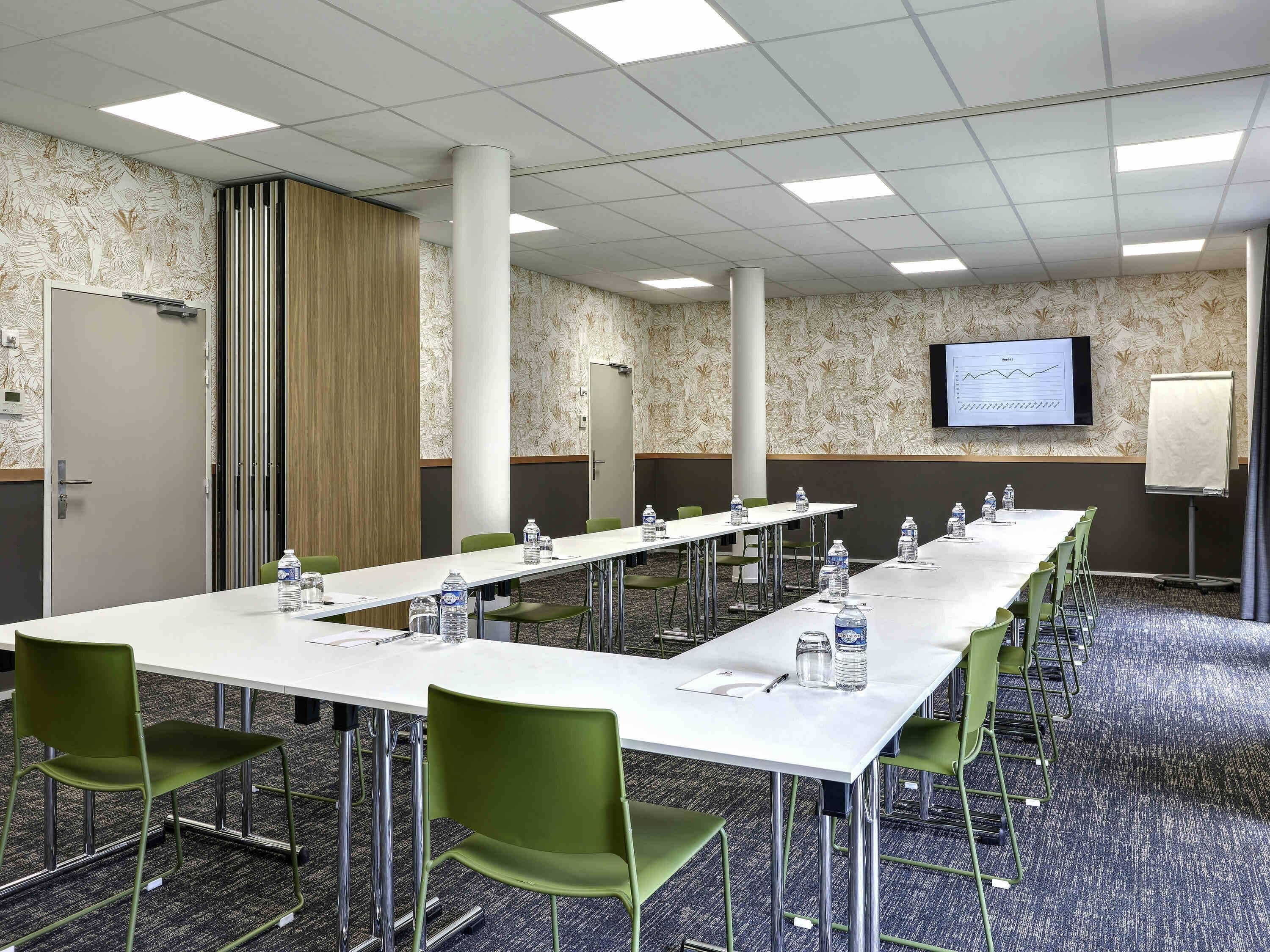 A conference room with long tables, green chairs, water bottles, a projector screen, and a whiteboard.