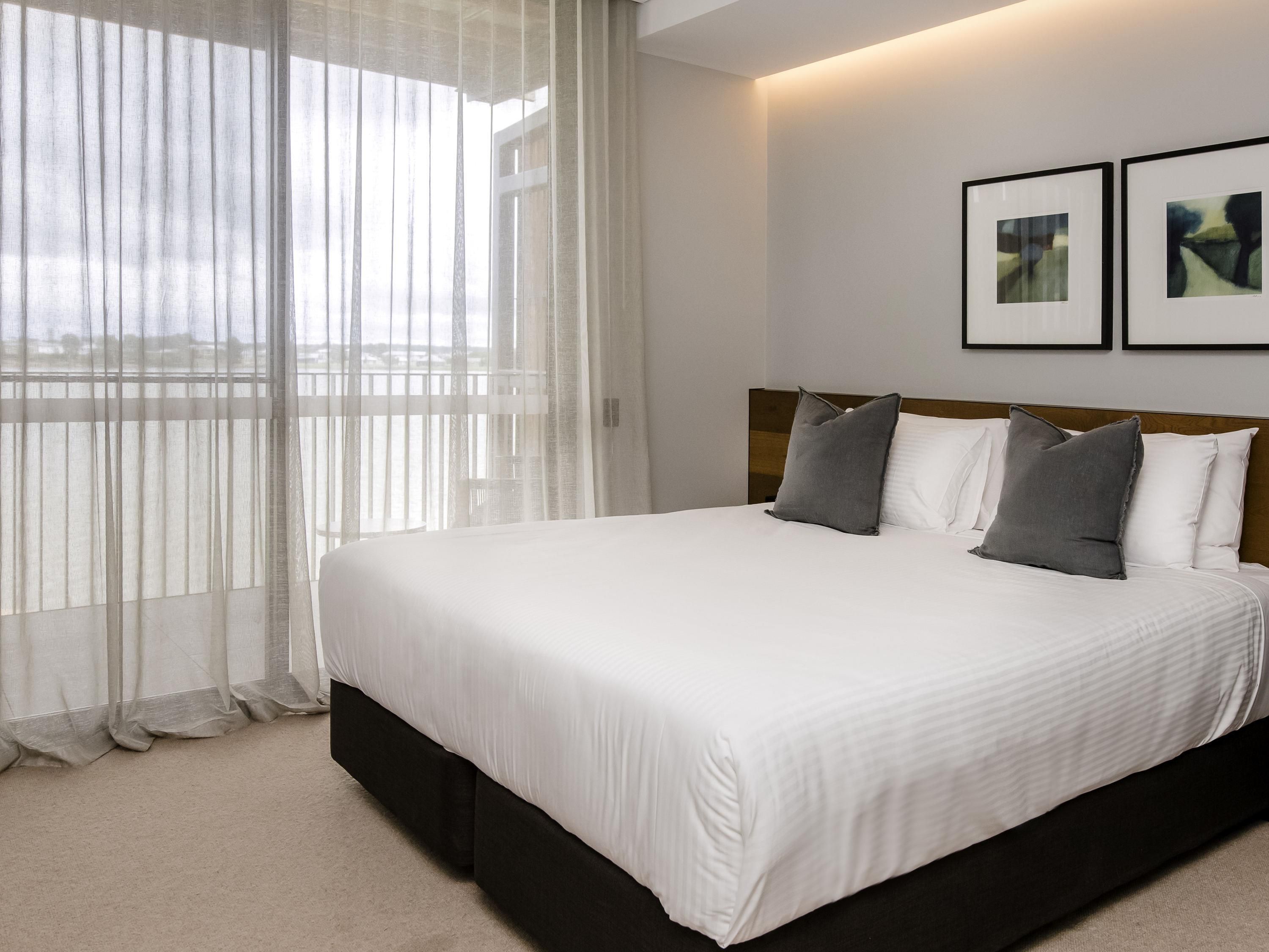 A neatly made bed with white bedding and two gray pillows, a wooden headboard, and sheer curtains on a window.
