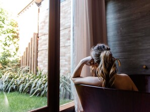 A person sitting in a bathtub with a window view of a brick wall and greenery outside. - The Sebel Yarrawonga Silverwoods (Yarrawonga)