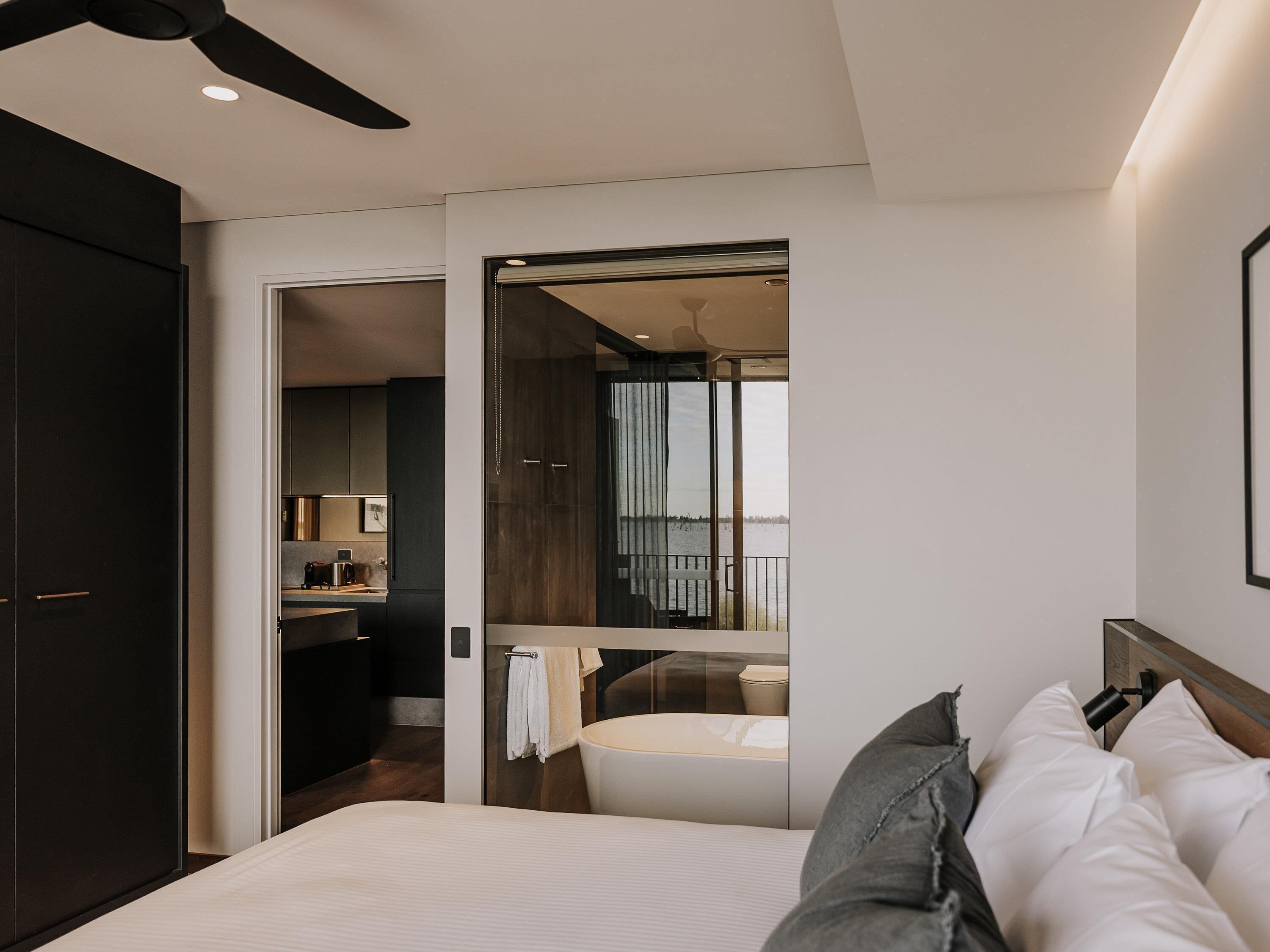A modern hotel room with a bed, a bathroom, a kitchenette, and a balcony with a view.