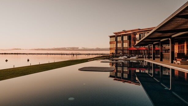 Un edificio moderno con piscina que refleja el cielo, una zona con césped y vistas al agua.