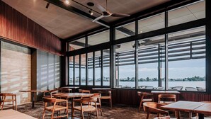Interior de restaurante con mesas y sillas de madera, ventanas amplias con vista a un cuerpo de agua y un ventilador de techo.