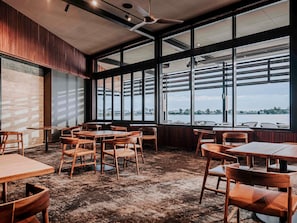Interior de restaurante con mesas y sillas de madera, ventanas amplias con vista a un cuerpo de agua y un ventilador de techo.