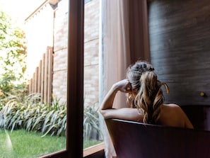 A person sitting in a bathtub with a window view of a brick wall and greenery outside. - The Sebel Yarrawonga Silverwoods (Yarrawonga)
