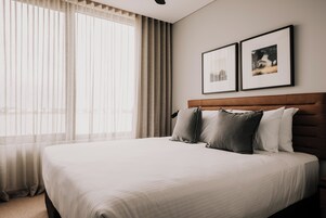 Una cama bien hecha con cabecera, un ventanal con cortinas translúcidas y dos cuadros en la pared.