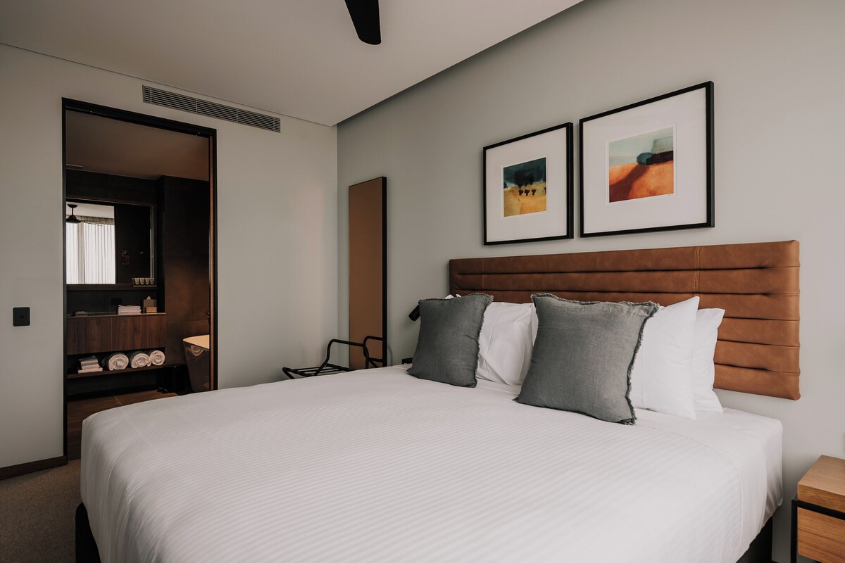 Habitación de hotel con una cama grande, cabecera de madera, dos cuadros enmarcados en la pared, una mesita de noche y un armario con toallas.