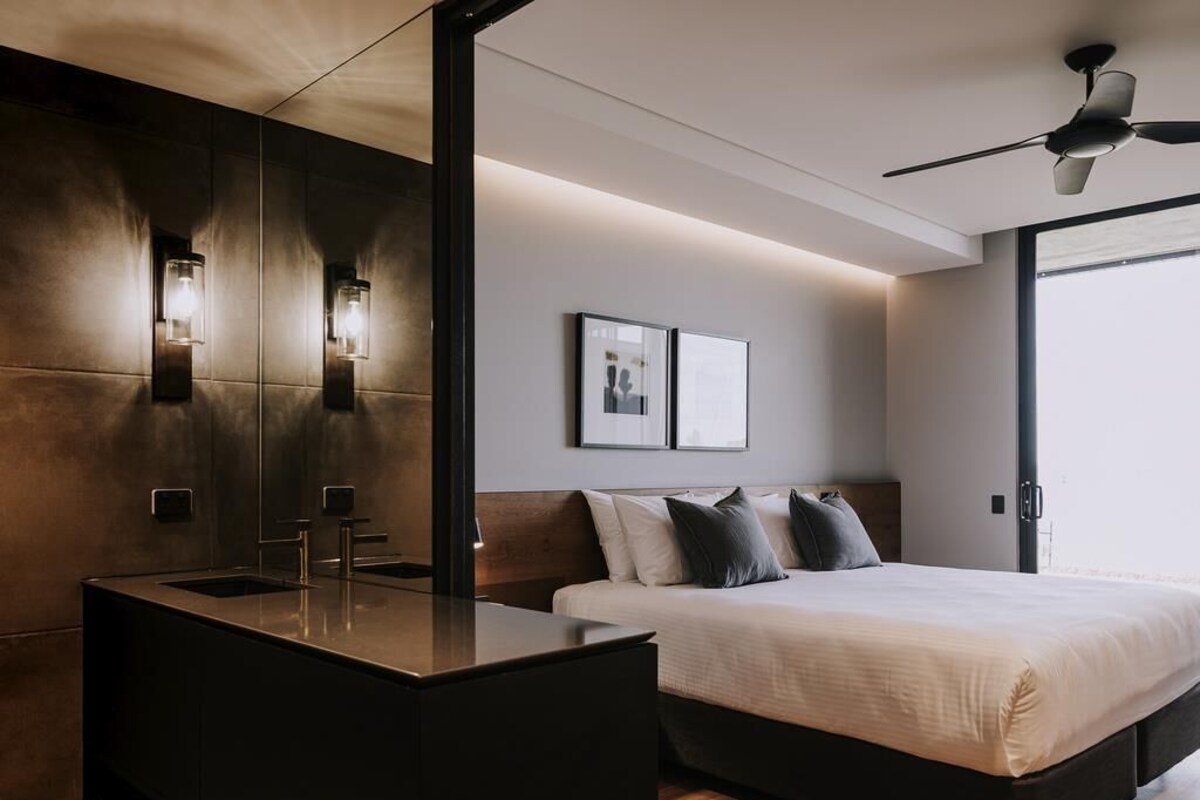 Habitación de hotel moderna con una cama grande, una mesita de noche, tocador empotrado con espejo e iluminación en la pared.