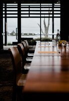 Un restaurante con una larga mesa de madera y sillas de cuero, grandes ventanales con vistas al agua y edificios, y una botella y un cuenco sobre la mesa.