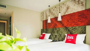 Une chambre d’hôtel avec deux lits, du linge de lit blanc, des oreillers rouges ornés du logo « ibis », une tête de lit en marbre et des lampes suspendues.