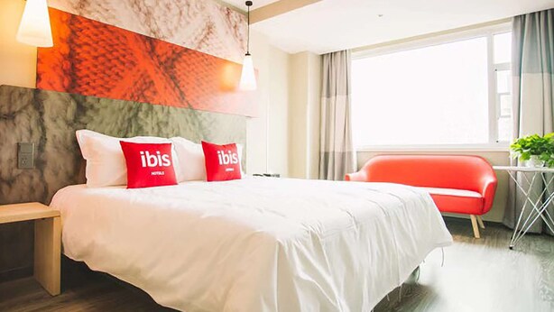 Une chambre d’hôtel avec un lit, une chaise rouge, une petite table avec une plante et une grande fenêtre.