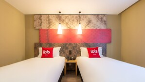 Een hotelkamer met twee bedden, elk met een rode ibis kussen, een houten nachtkastje met een telefoon en een wand met een marmerpatroon boven de bedden.