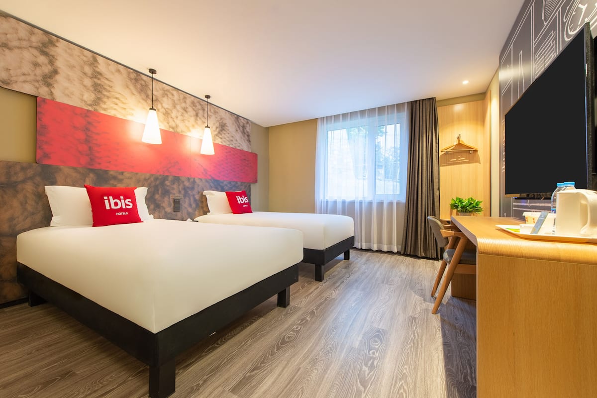 Ein Hotelzimmer mit zwei Betten, einem Schreibtisch, einem Fernseher und einem großen Fenster mit Vorhängen.