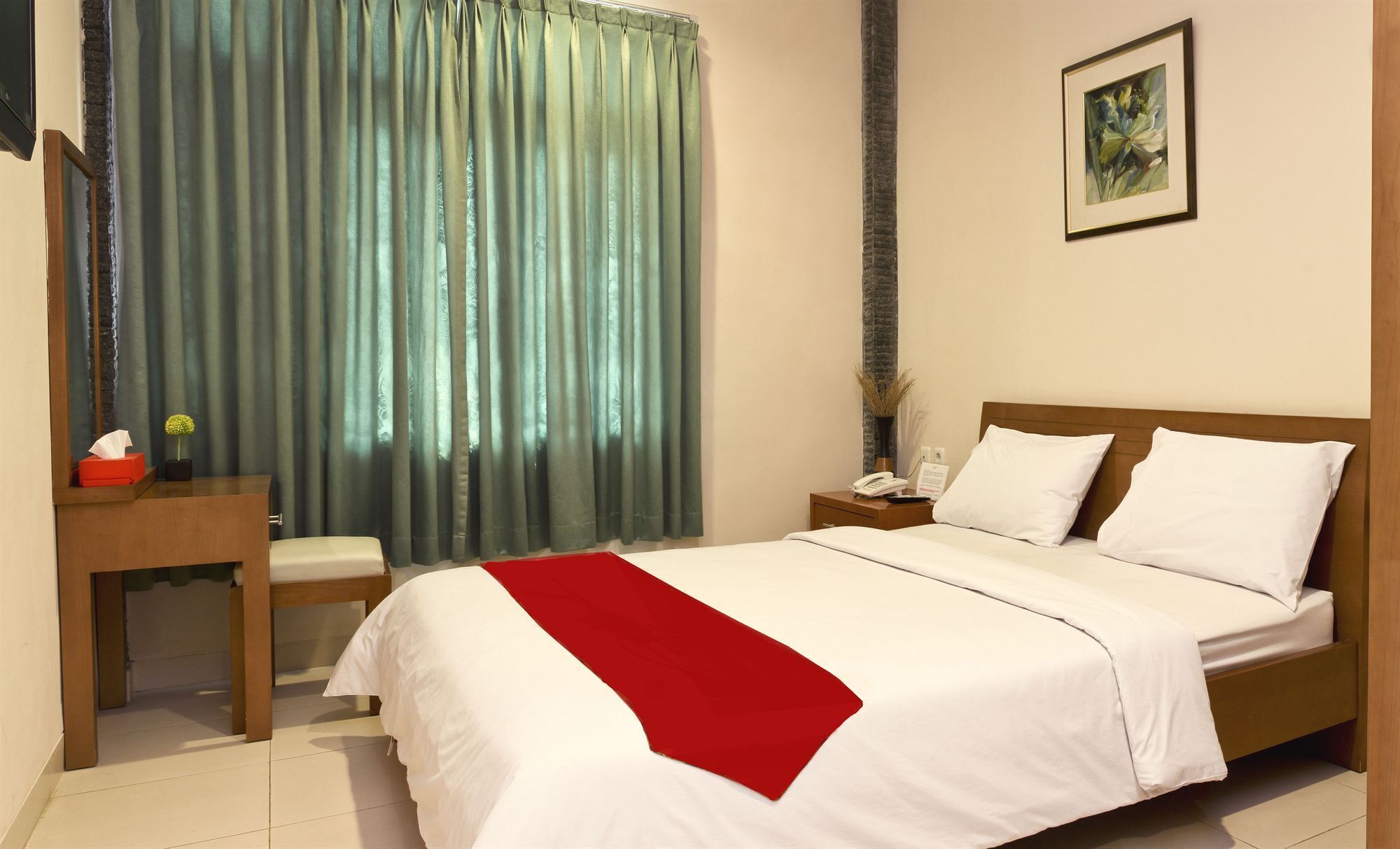 A hotel room with a bed, a desk, a chair, a nightstand, a telephone, a vase with flowers, and a framed picture on the wall.