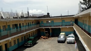 A multi-story building with a central courtyard. There are three cars parked in the courtyard, a green truck, and a white van. The building has a flat roof with some equipment on top. - Heshima Lodge (Naivasha)