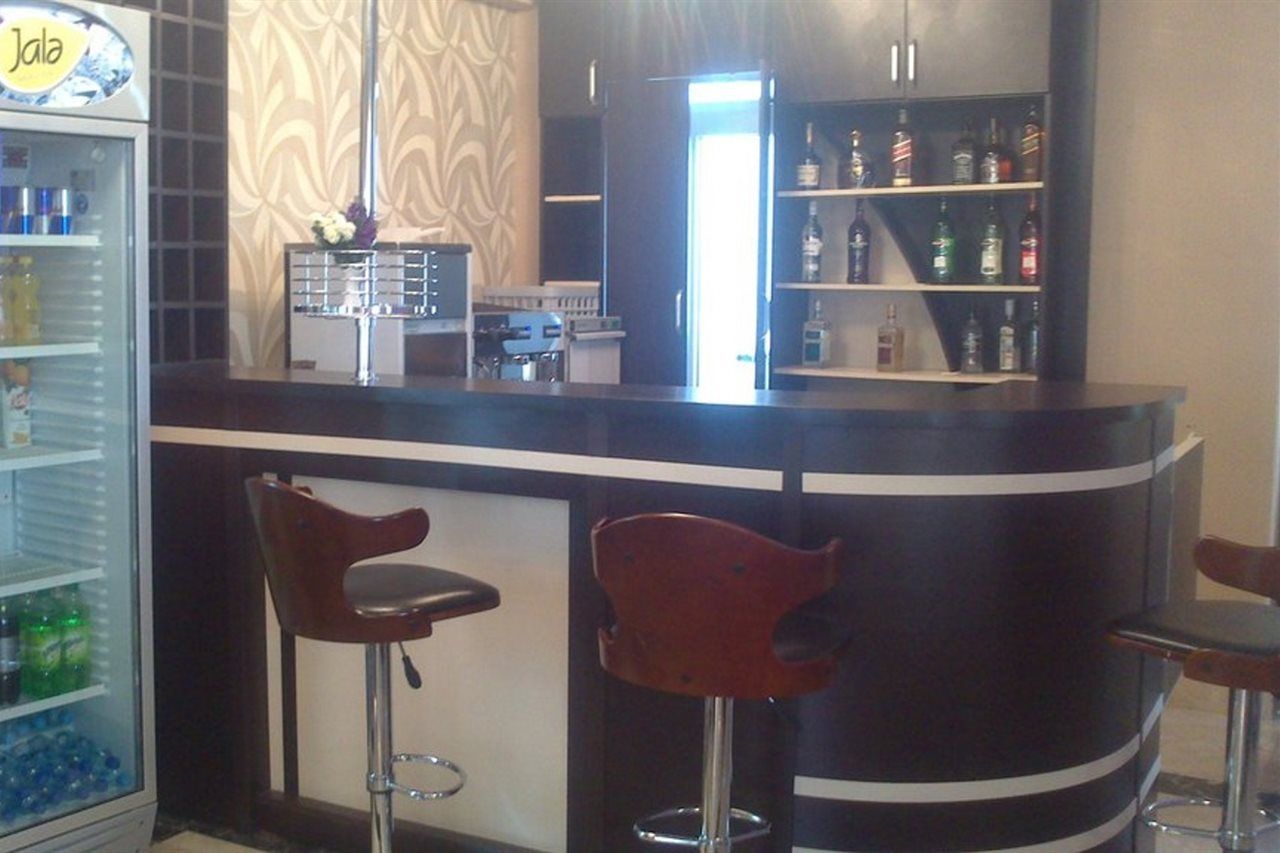 A bar area with a curved counter, two stools, a refrigerator stocked with beverages, and a shelf with liquor bottles.