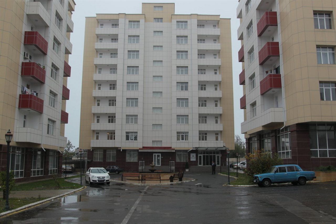 A multi-story residential building with a central entrance, a parking area with cars, and a small outdoor seating area.
