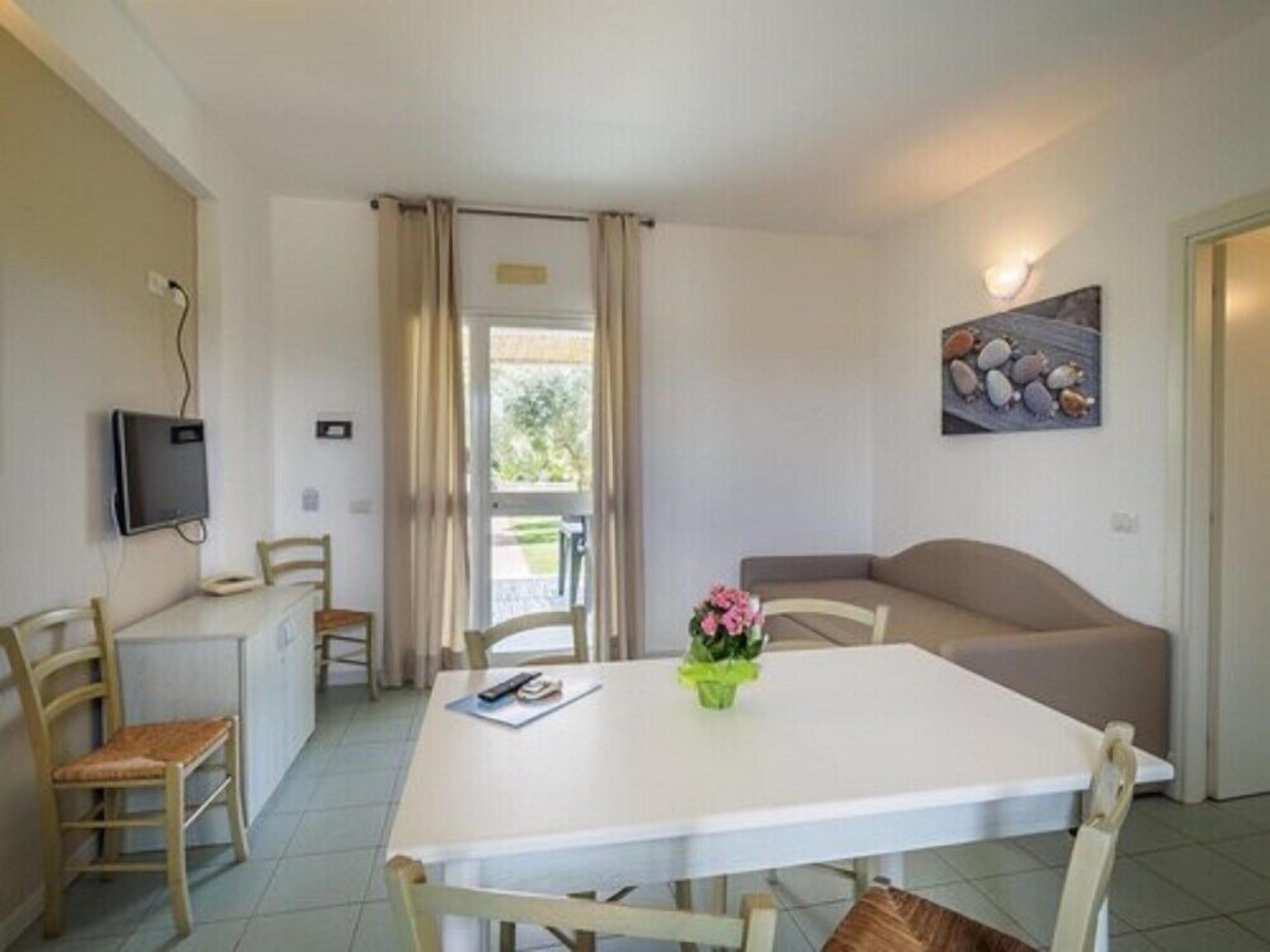 Une petite pièce bien éclairée, avec une table blanche, deux chaises, un canapé, un téléviseur fixé au mur et une porte donnant sur un espace extérieur.