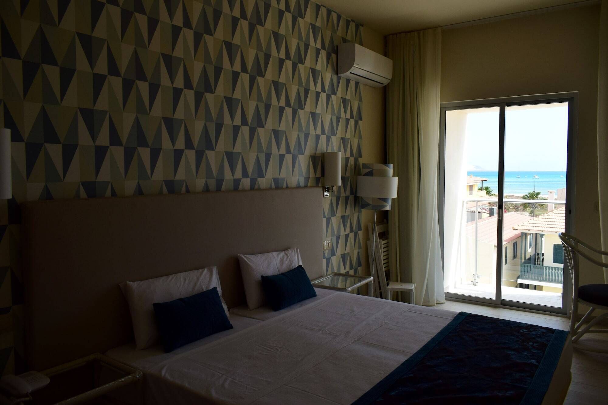 Une chambre d’hôtel avec un grand lit, deux oreillers, une table de chevet, une lampe et une vue sur l’océan par une porte-fenêtre coulissante.