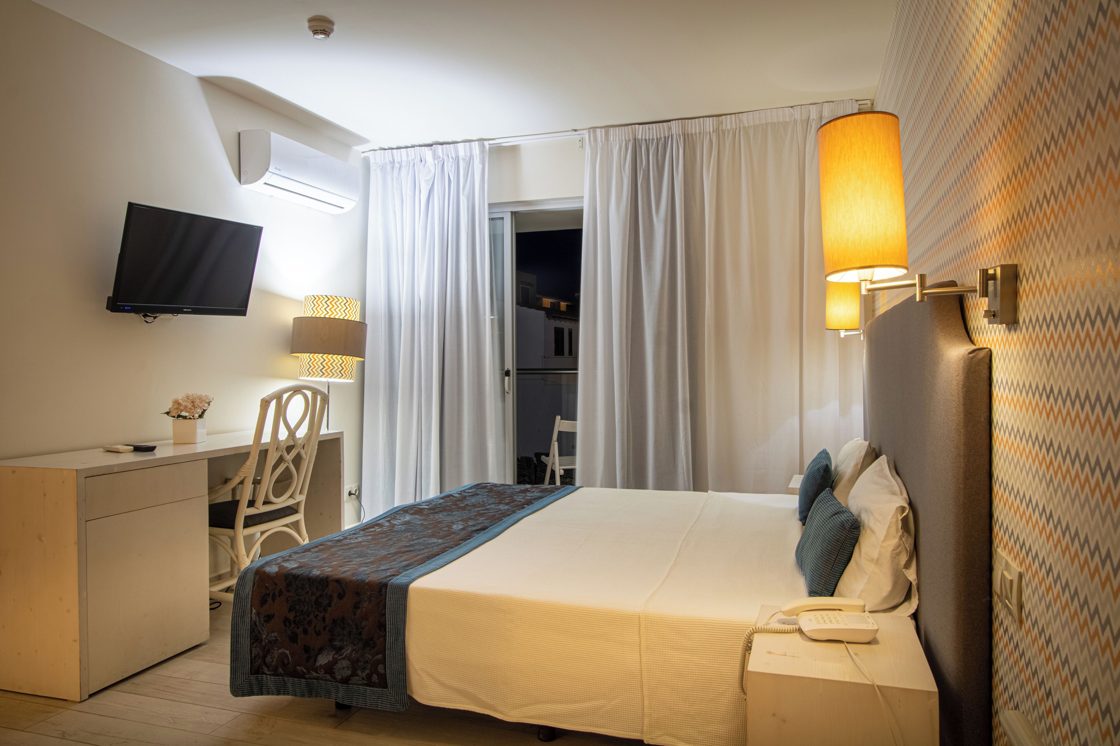 A hotel room with a bed, a desk, a chair, a television, and a lamp.
