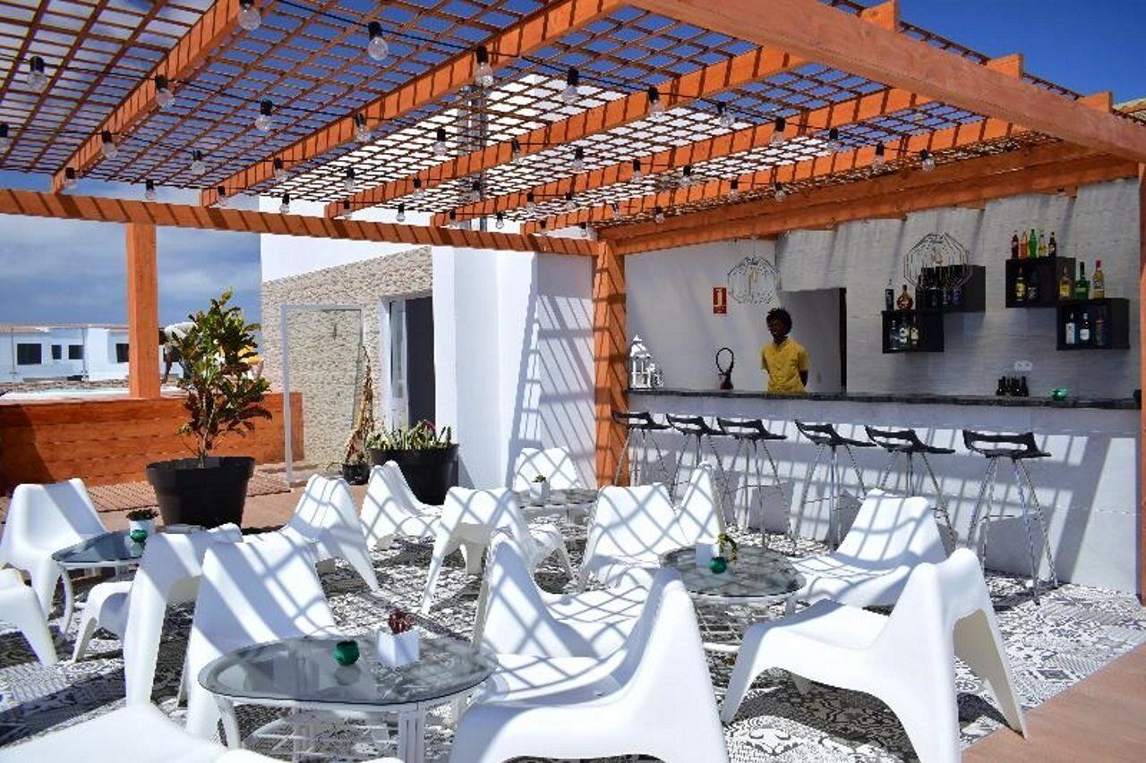 An outdoor patio with white plastic chairs and tables, a bar area with shelves of bottles, and a person in the background.