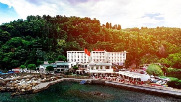 Un hotel frente al mar con una construcción grande, un muelle con barcos y un paisaje boscoso al fondo.